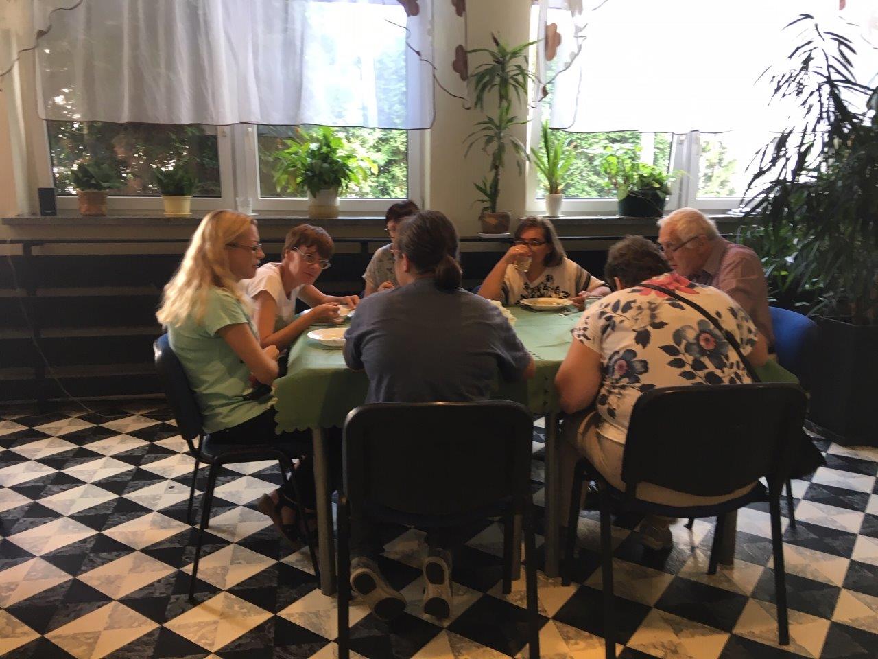 Zjazdowicze podczas posiłku w jadalni z charakterystyczną biało-czarną podłogą w stylu szachownicy, siedzący przy kilkuosobowym stole przy oknie z gładkimi białymi firankami