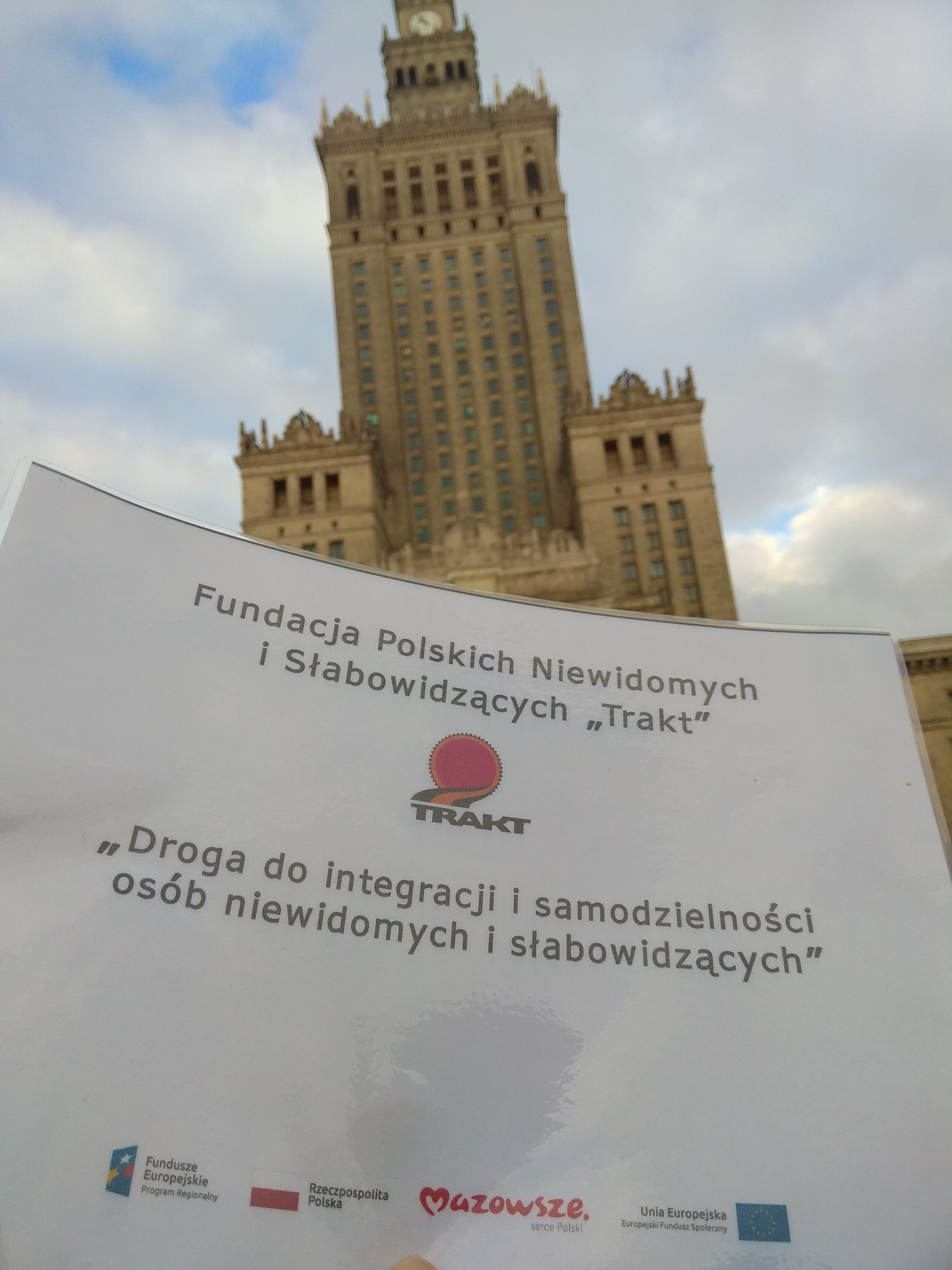 Pałac Kultury i Nauki w Warszawie – wysoki, strzelisty budynek w jasnej kolorystyce z wieloma oknami , a pod nim wkomponowany plakat projektu unijnego.