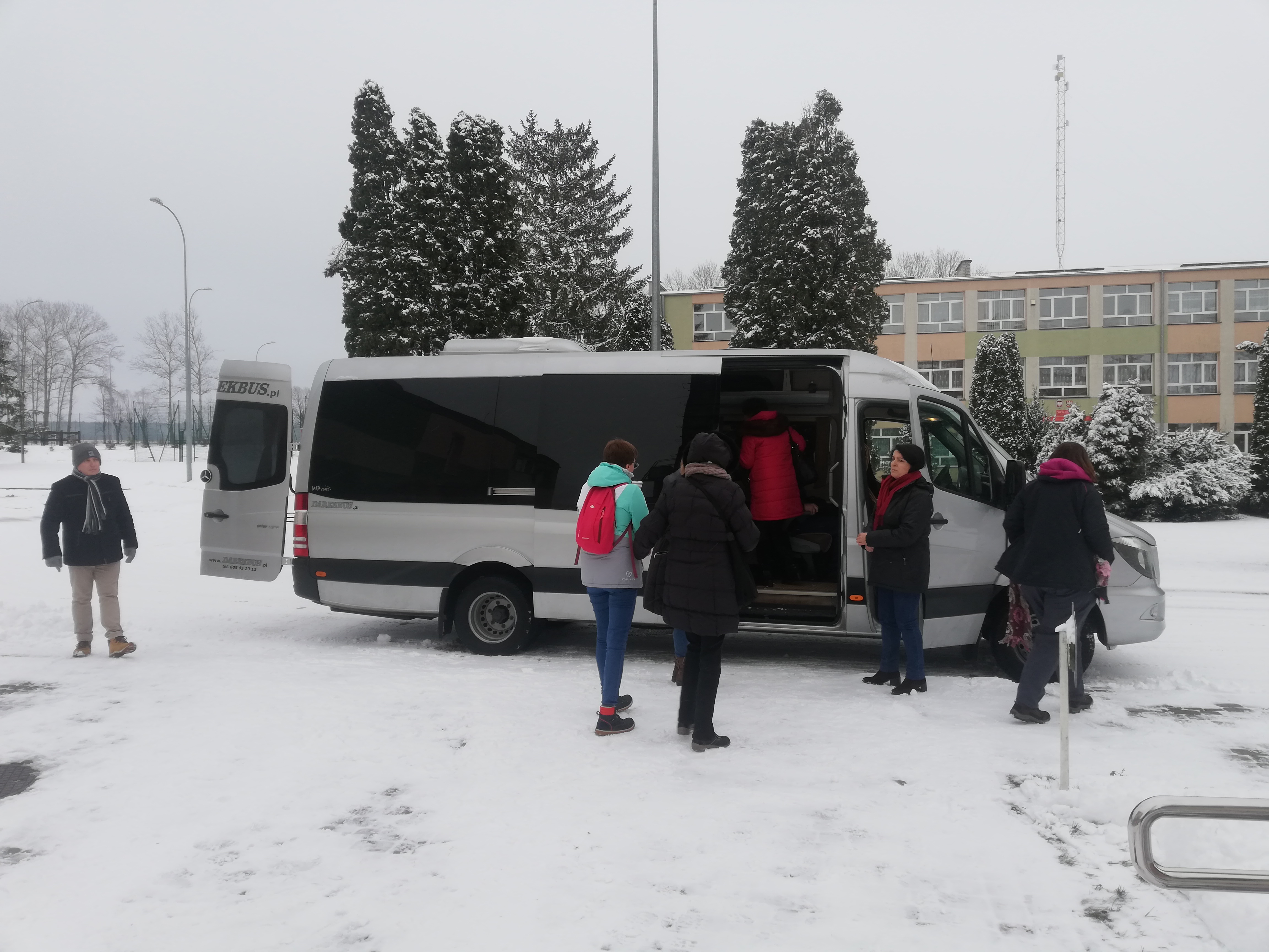 Przed odjazdem –biały bus z otwartymi drzwiami i tylnym bagażnikiem, przy nim kilkoro uczestników na chwilę przed wyjazdem z Krzyżewa, ziemia pokryta śniegiem.